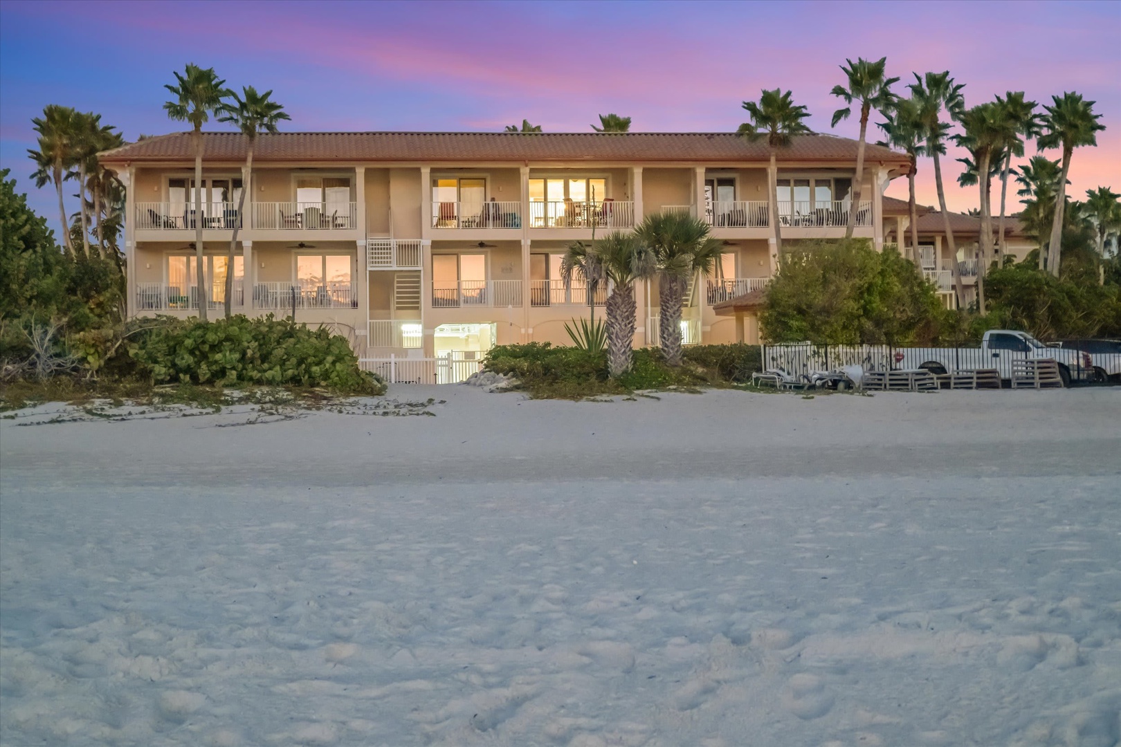 A charming beachfront property nestled among tropical palms, photographed at twilight as warm lights illuminate the balconies and the sky glows in soft hues of pink and blue.