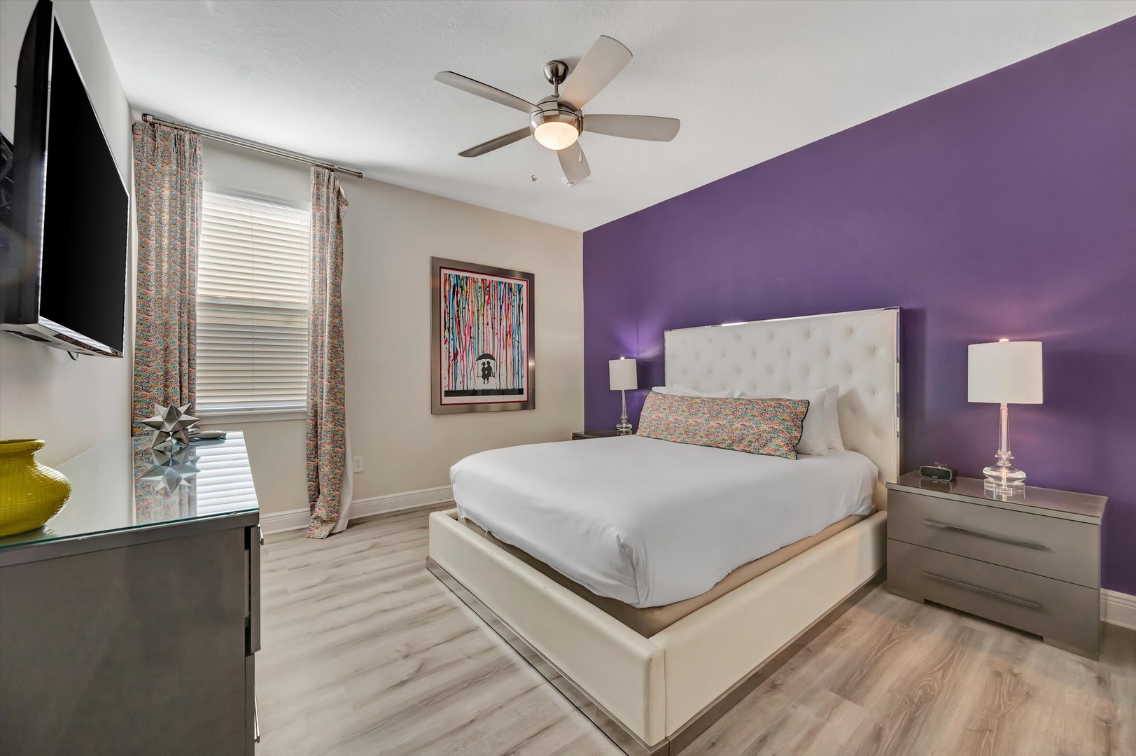 Unwind in your stylish bedroom featuring a plush bed, modern décor, and a calming purple accent wall—perfect for peaceful nights after exploring the area.