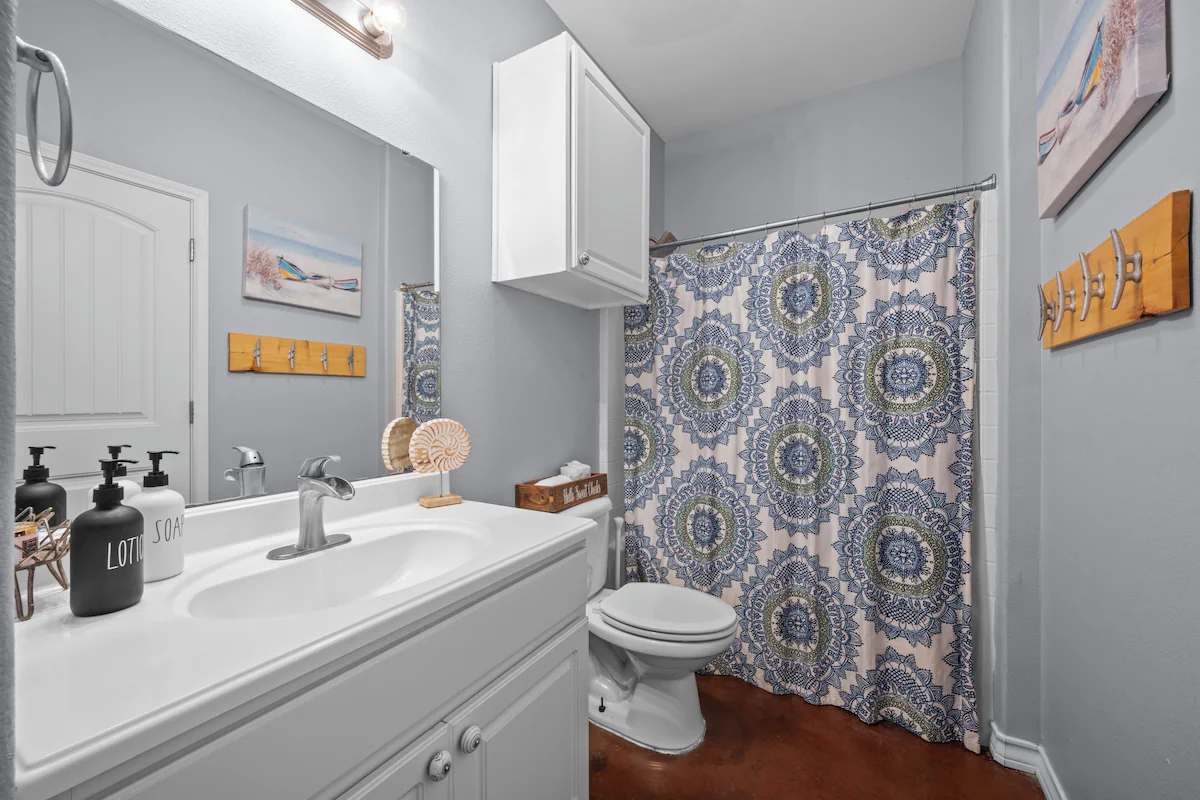 Step into this fresh, coastal-inspired bathroom where beach-themed accents create a relaxed vacation vibe. The soft gray walls and elegant medallion shower curtain add charm, while thoughtful touches make you feel right at home.