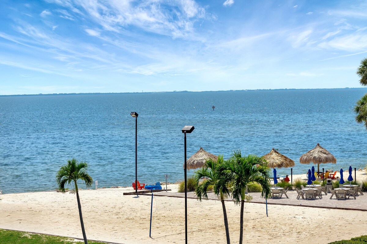 A private beachfront retreat stretches along crystal-blue waters, with swaying palms and thatched umbrellas dotting pristine white sand. The panoramic bay view sets the scene for endless sunny days by the shore.