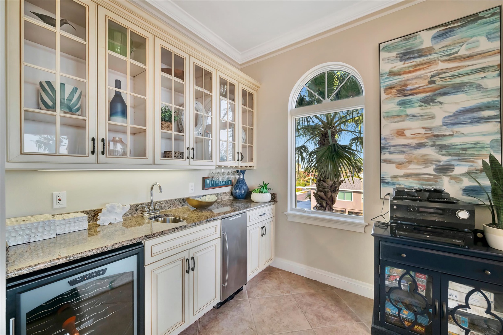 Your butler's pantry features granite counters, glass-front cabinetry, and a sink for seamless entertaining, all bathed in light from an arched window overlooking swaying palms.