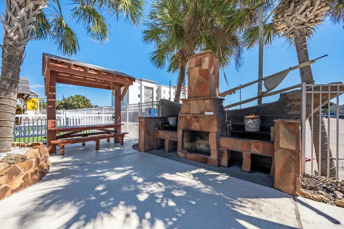 An outdoor BBQ area featuring brick grills and a shaded pergola, surrounded by palm trees and sunshine—perfect for enjoying a cookout with your group.