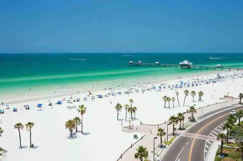 This stunning coastal area features pristine white sand beaches and emerald waters, with a nearby pier extending into the Gulf. Palm trees line the beachfront promenade, part of the vibrant surrounding neighborhood.