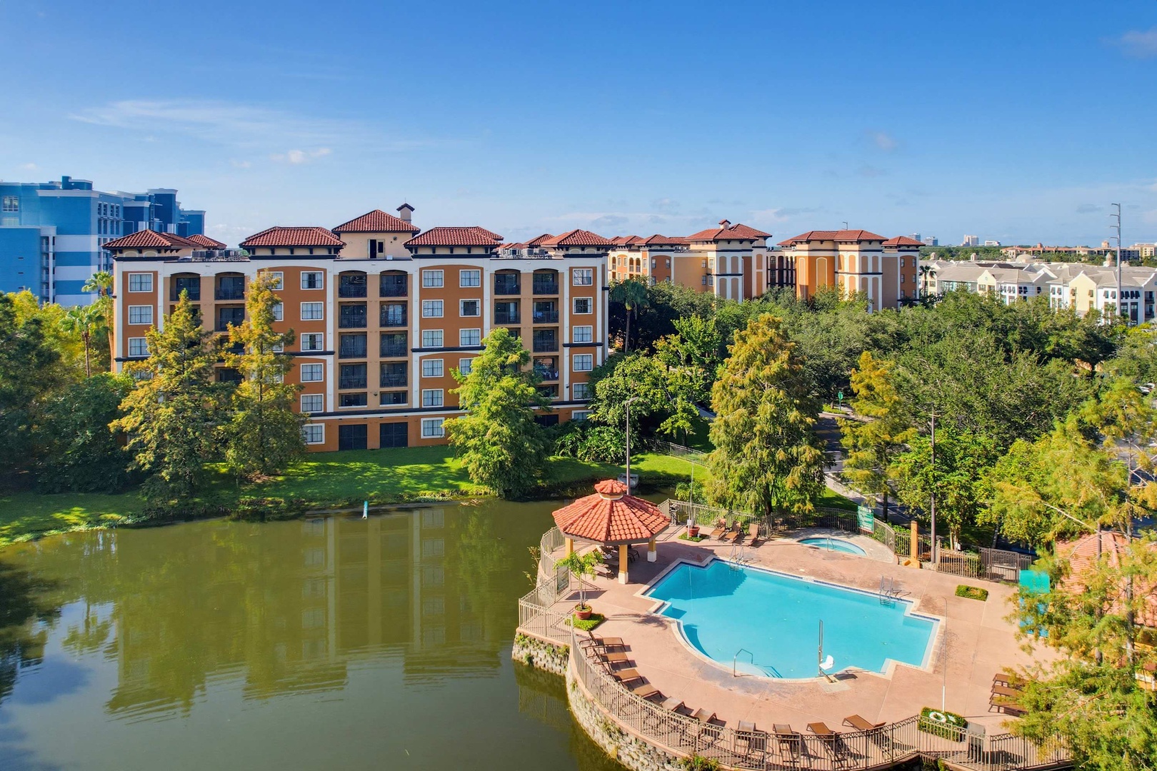 Aerial view of a lakeside resort property featuring Mediterranean-style buildings, lush mature trees, and a sparkling outdoor pool surrounded by sun loungers.