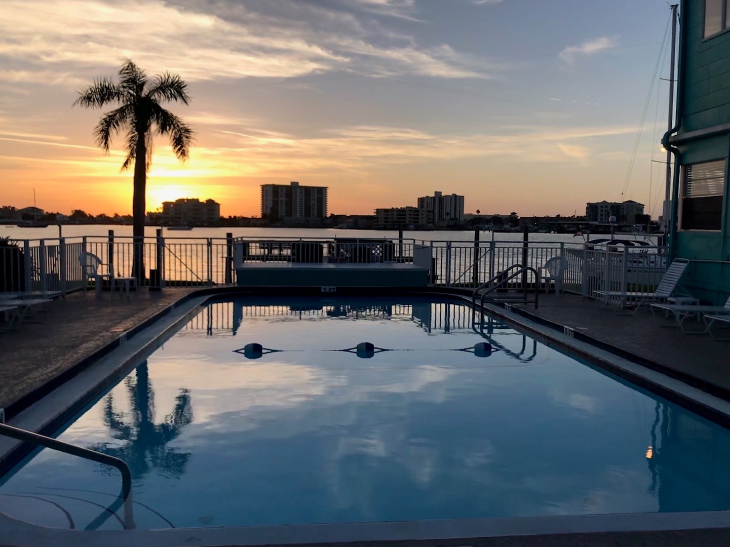 A stunning coastal sunset paints the sky over the waterfront pool, where golden light reflects off calm waters and silhouetted palms frame the horizon.
