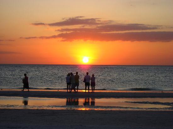 Golden sunset paints the sky over calm waters as silhouettes gather on the beach, marking the perfect end to a coastal day in this peaceful waterfront setting.