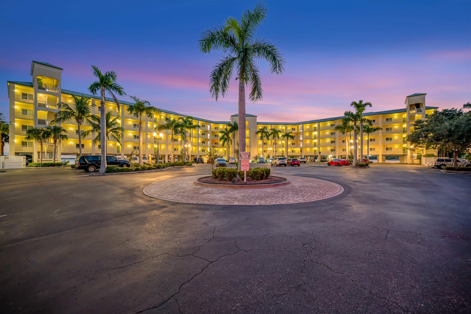 The property stands beneath a breathtaking twilight sky, where pink and purple hues blend into deepening blue, while palm trees frame the welcoming entrance and warm lights glow from every level.