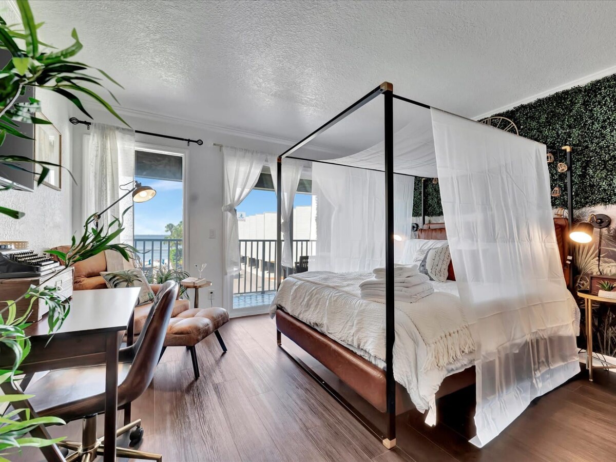 Drift into restful sleep in your canopy bed draped in soft white linens, with the ocean just beyond the balcony doors and lush greenery framing your peaceful retreat.