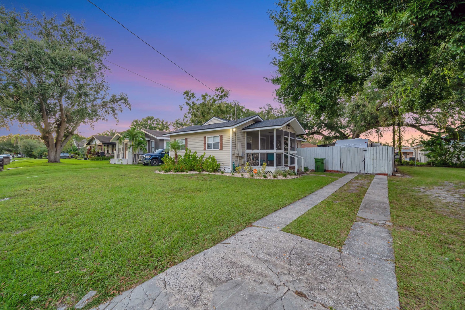 The property sits in a peaceful residential neighborhood, framed by mature trees and green lawns, with colorful skies marking the transition between day and night.