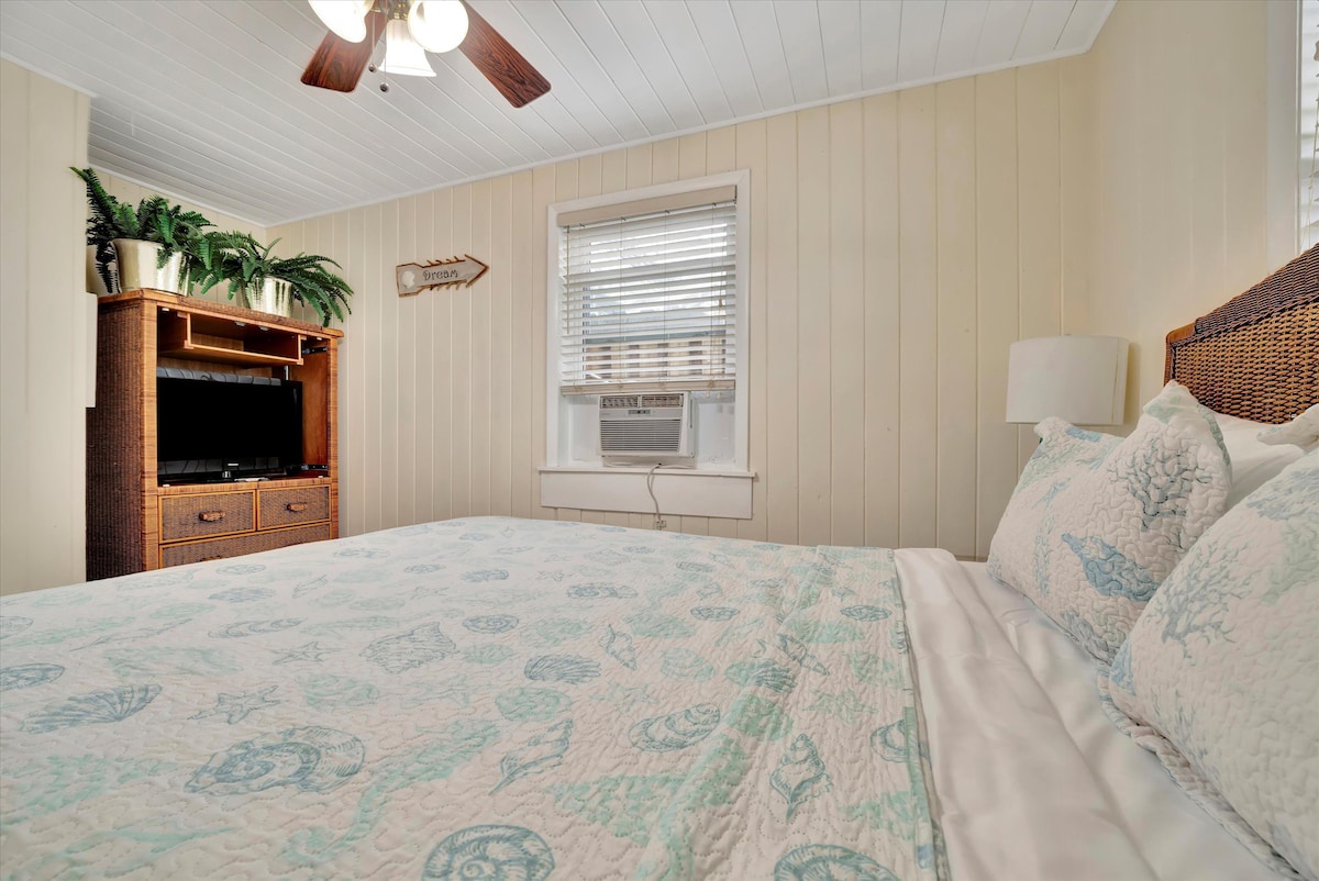 Serene coastal bedroom with soft ocean-inspired linens, warm wood accents, and wicker furniture. A ceiling fan keeps things breezy while gentle lighting creates the perfect retreat for restful nights.