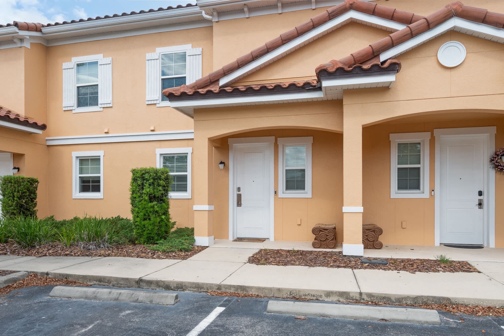 Two-story townhome with warm stucco exterior, terracotta tile roof, white shuttered windows, and covered entryway. The property features landscaped grounds with greenery and decorative columns at each front door.
