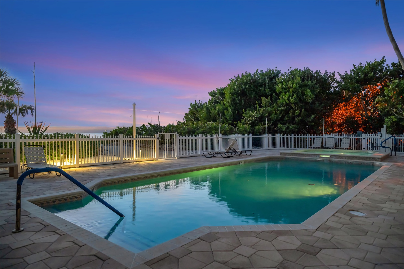 A tranquil evening settles over the pool area as gentle lighting illuminates the water. Colorful skies transition from blue to soft pinks, while swaying palms and lush tropical greenery frame your private oasis.