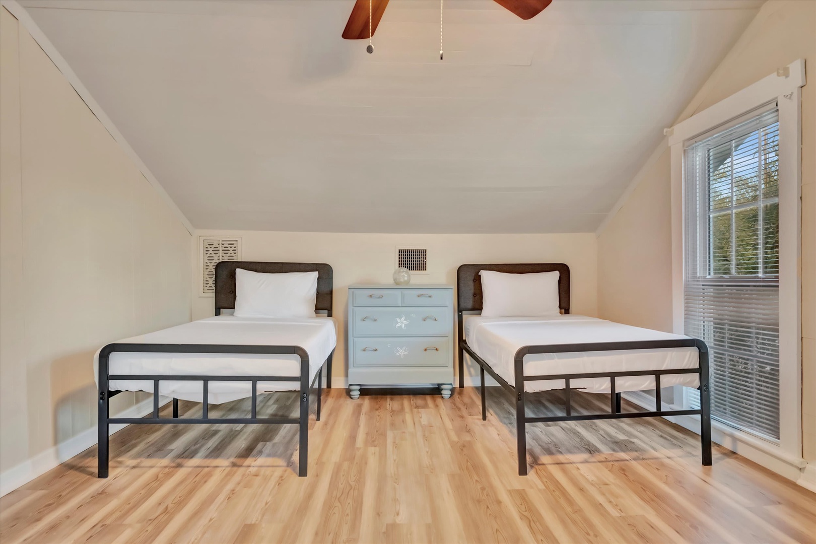 Your cozy bedroom features two comfortable twin beds with fresh linens, warmed by a ceiling fan above and natural light streaming through the window.