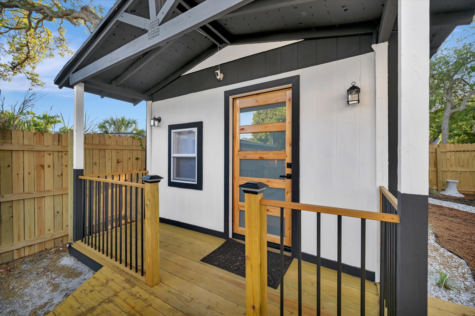 Modern cottage exterior featuring a sleek black and white facade with warm wood accents, covered porch, and surrounded by natural landscaping with mature trees.