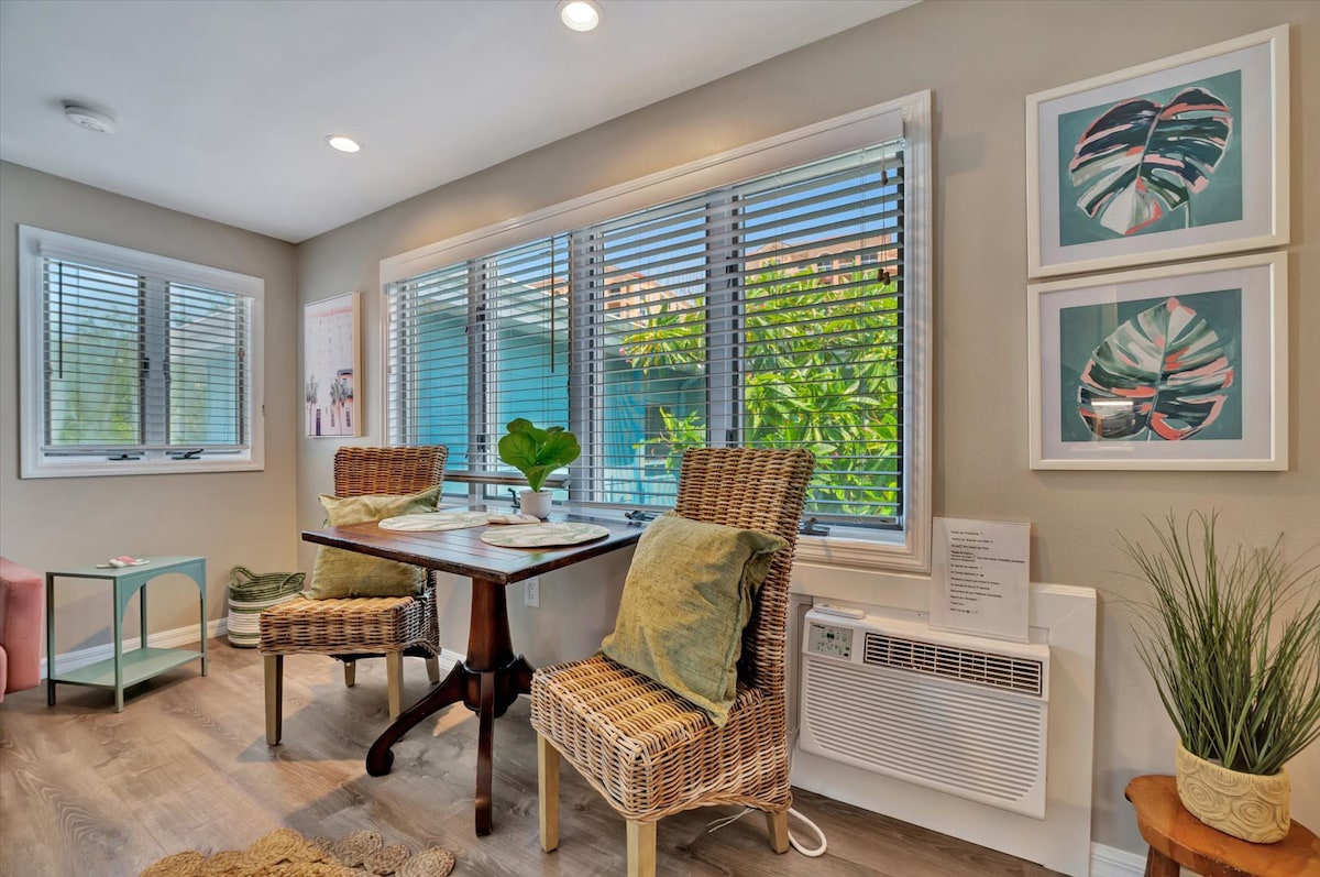 Cozy breakfast nook with natural light, tropical artwork, woven chairs, and air conditioning for year-round comfort.
