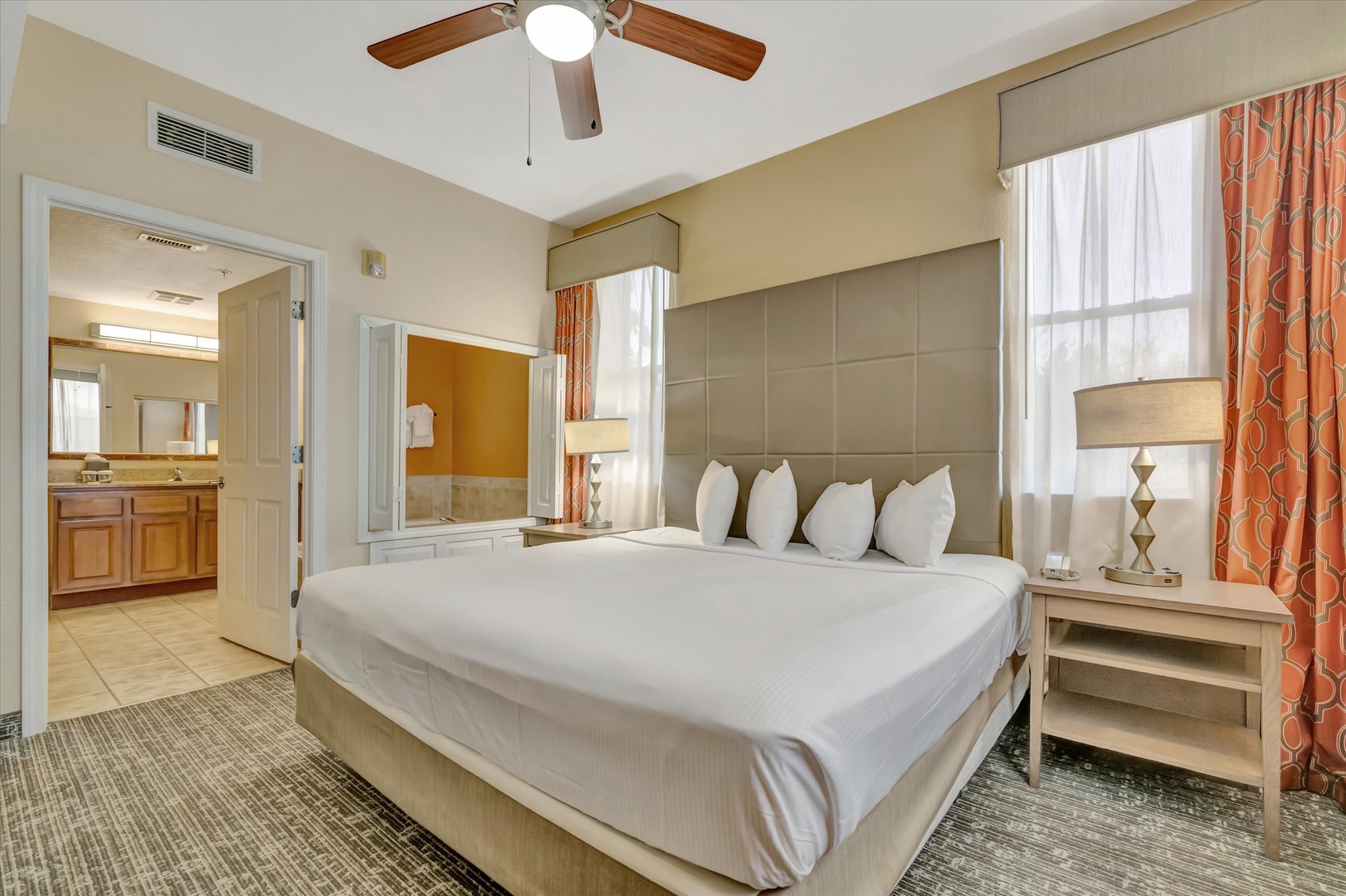 Unwind in your modern bedroom featuring a plush king bed, stylish padded headboard, and elegant furnishings. A ceiling fan keeps you comfortable, while warm coral accents add a welcoming touch to your private retreat.