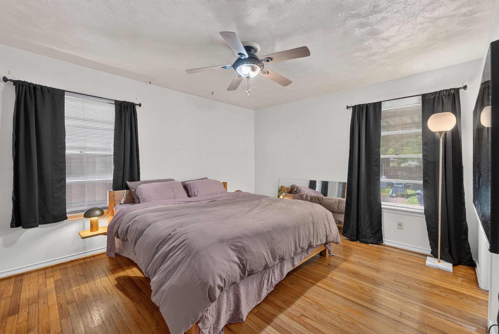 Your peaceful bedroom features warm hardwood floors, crisp white walls, and dark curtains that frame natural light streaming through large windows—perfect for restful nights.