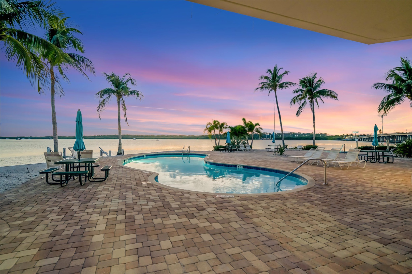 The property's waterfront pool area, framed by palm trees and surrounded by paver stone deck, overlooks tranquil bay waters at sunset—a peaceful setting for arrival and relaxation near the pier.