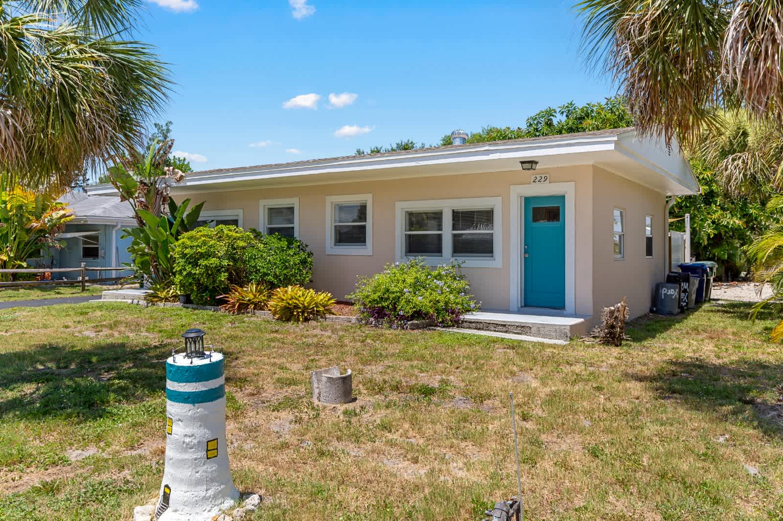 Charming coastal cottage featuring a bright turquoise door and surrounded by lush tropical landscaping, including palm trees and native plants, creating a welcoming retreat in a peaceful neighborhood.