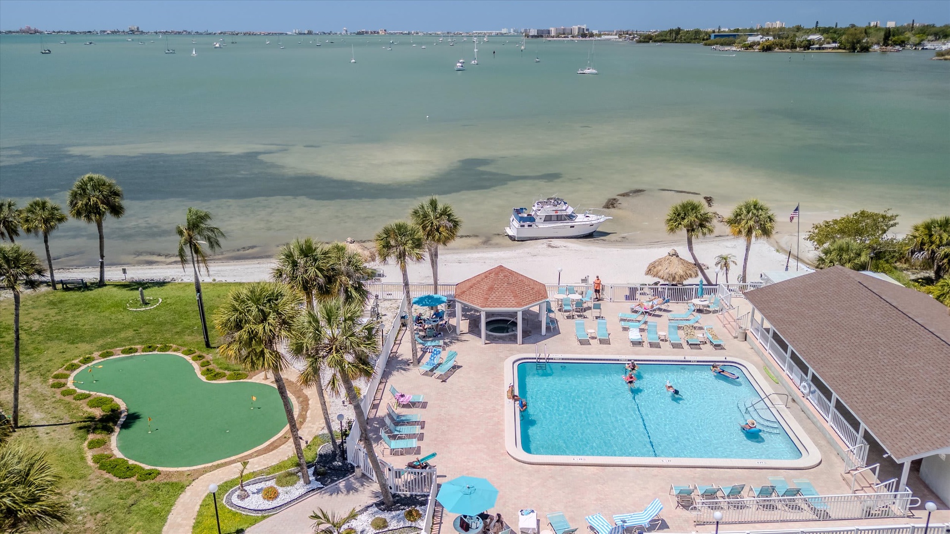This aerial view showcases the property's waterfront setting with direct beach access, a sparkling pool, mini-golf course, and palm-studded grounds along peaceful bay waters perfect for boating.