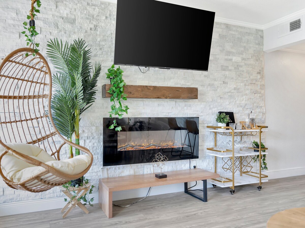 Relax in your bright, modern living space featuring a cozy electric fireplace, unique hanging chair, and stylish stone accent wall—perfect for unwinding after exploring the area.