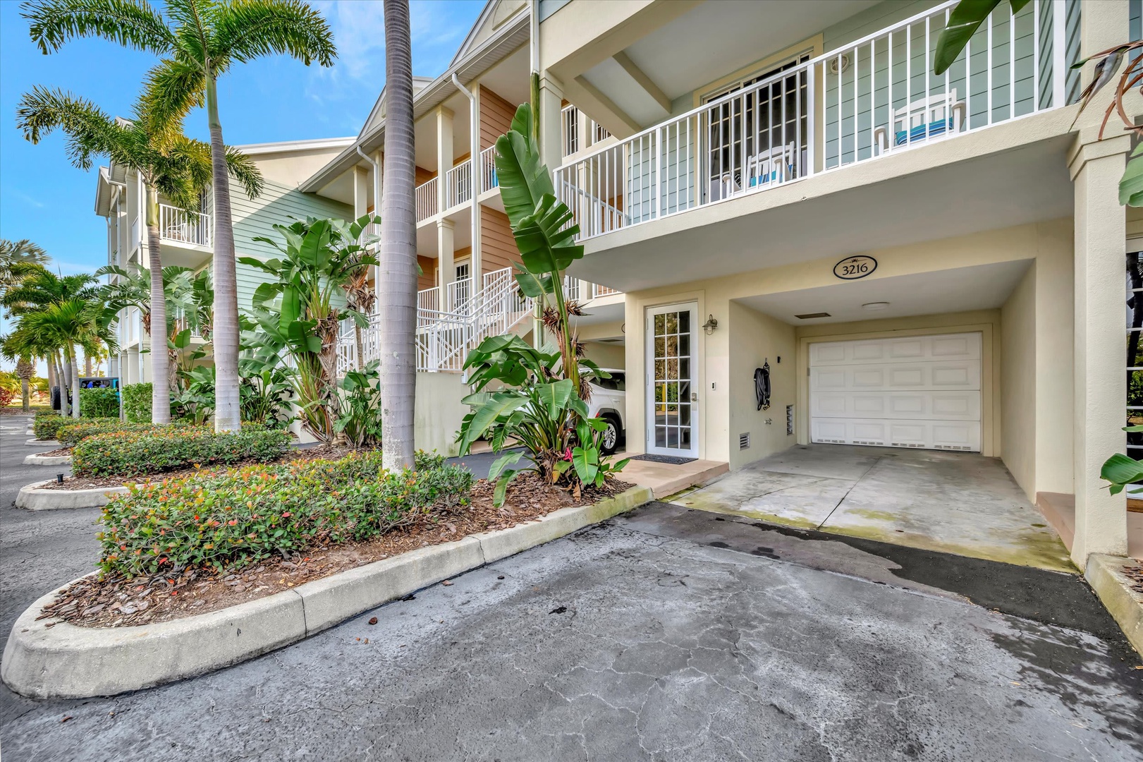 A tropical multi-unit building with lush palm trees and landscaped gardens marks the property entrance, featuring private garage parking beneath spacious balconies.