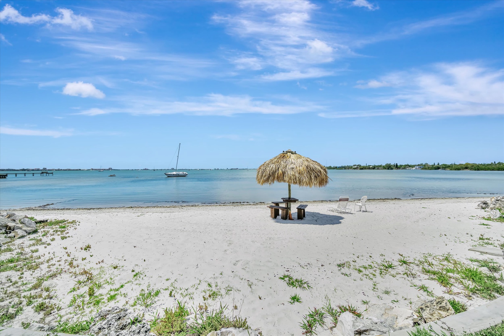 A private beach area with tiki-style shade and seating overlooking calm turquoise waters and distant sailboats.