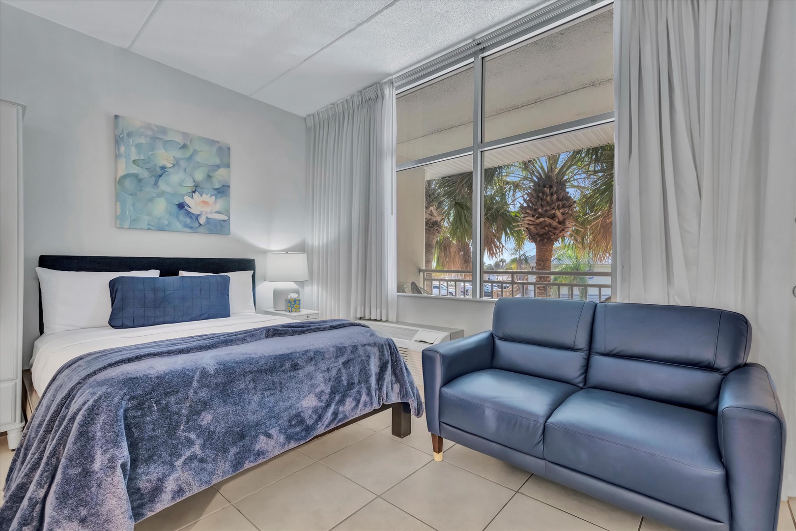 Settle into your serene bedroom, where a plush bed dressed in soft blues invites restful sleep. A stylish sofa provides extra lounging space, and the large window frames tropical palms outside, filling your room with natural light.
