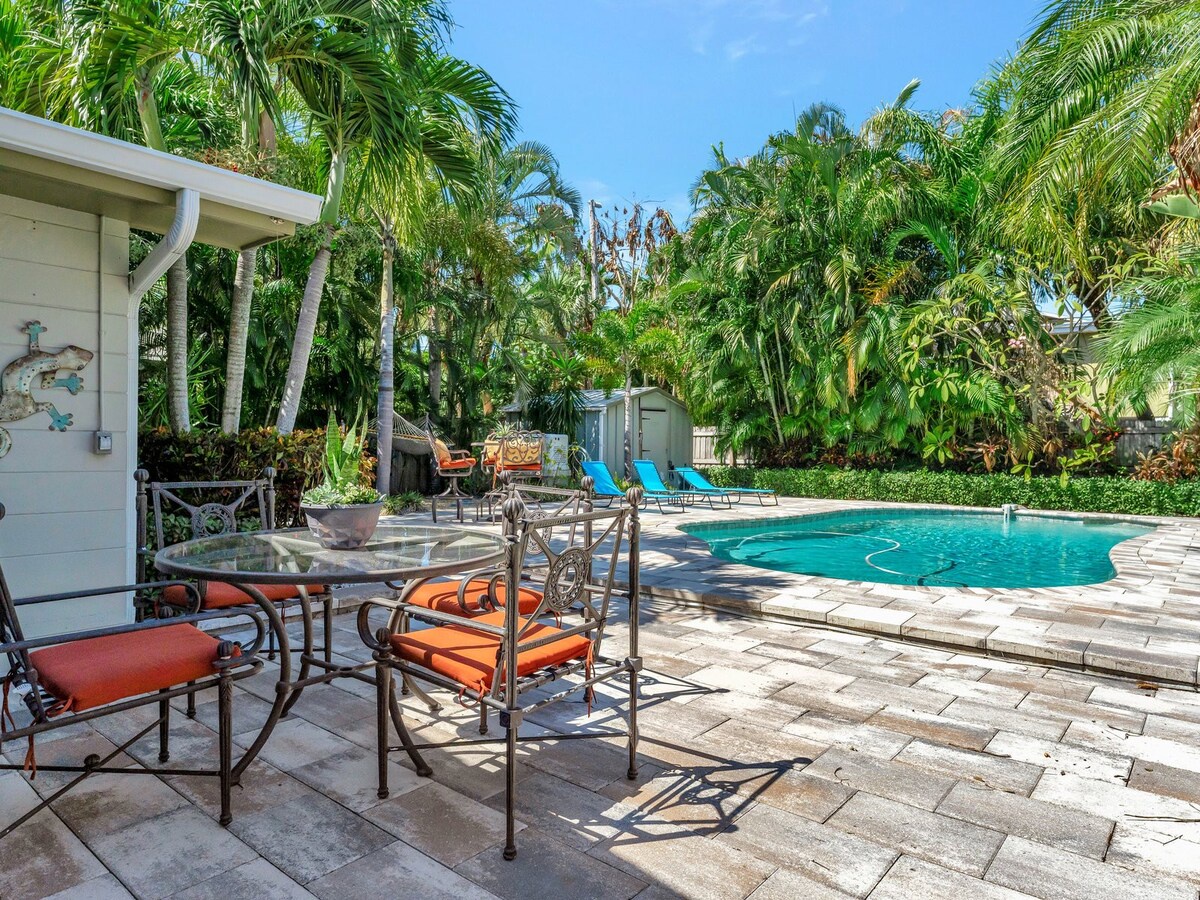 A sparkling pool surrounded by lush tropical palms and lounge chairs awaits, with shaded patio seating perfect for morning coffee or sunset cocktails in your private backyard oasis.