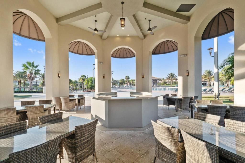 A welcoming open-air pavilion with elegant arched windows and wicker seating overlooks lush palm-lined surroundings and a sparkling pool in the distance.