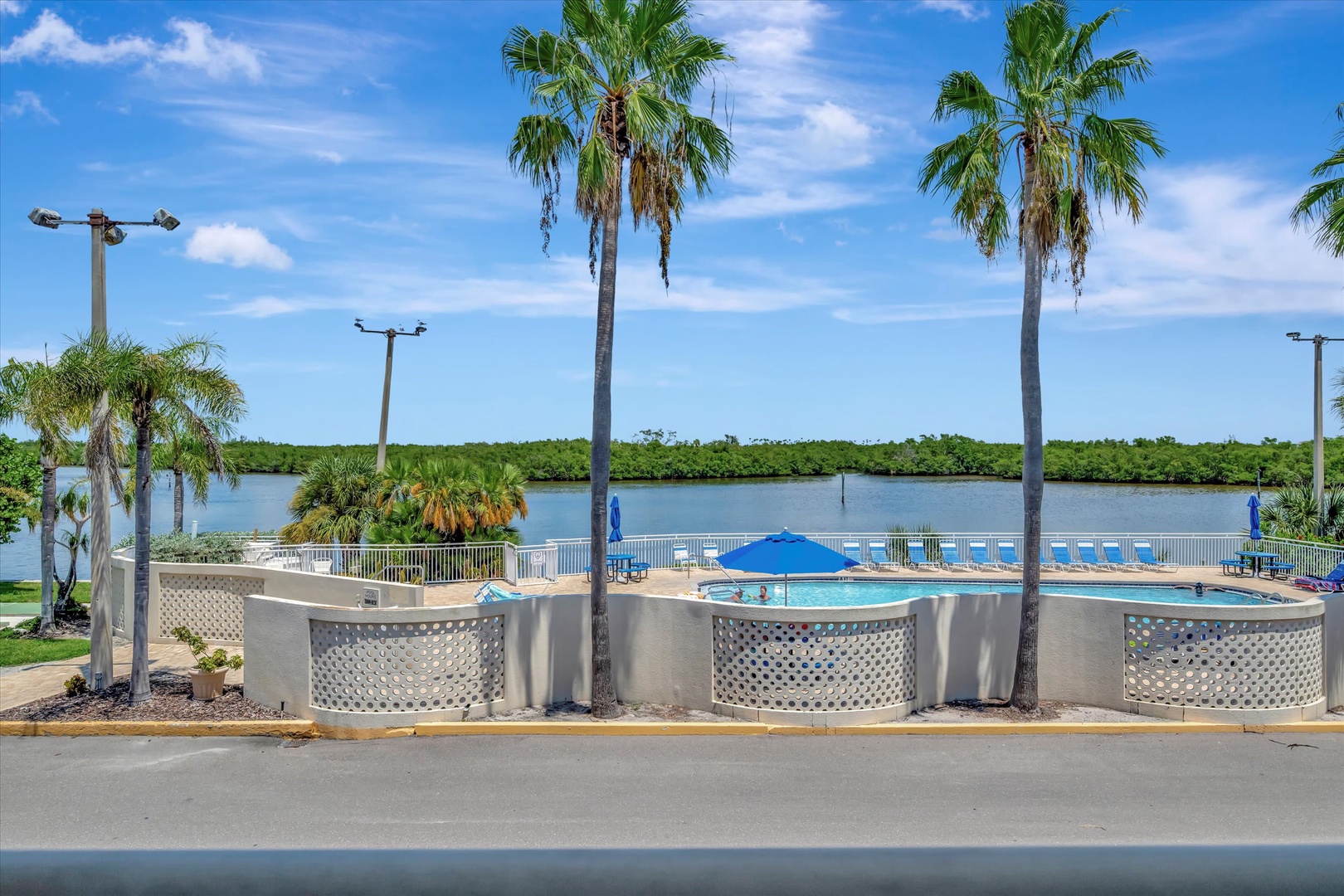 The property features a waterfront pool area surrounded by palm trees and overlooking a scenic river with lush mangroves beyond. Blue lounge chairs and umbrellas invite relaxation by the water's edge.