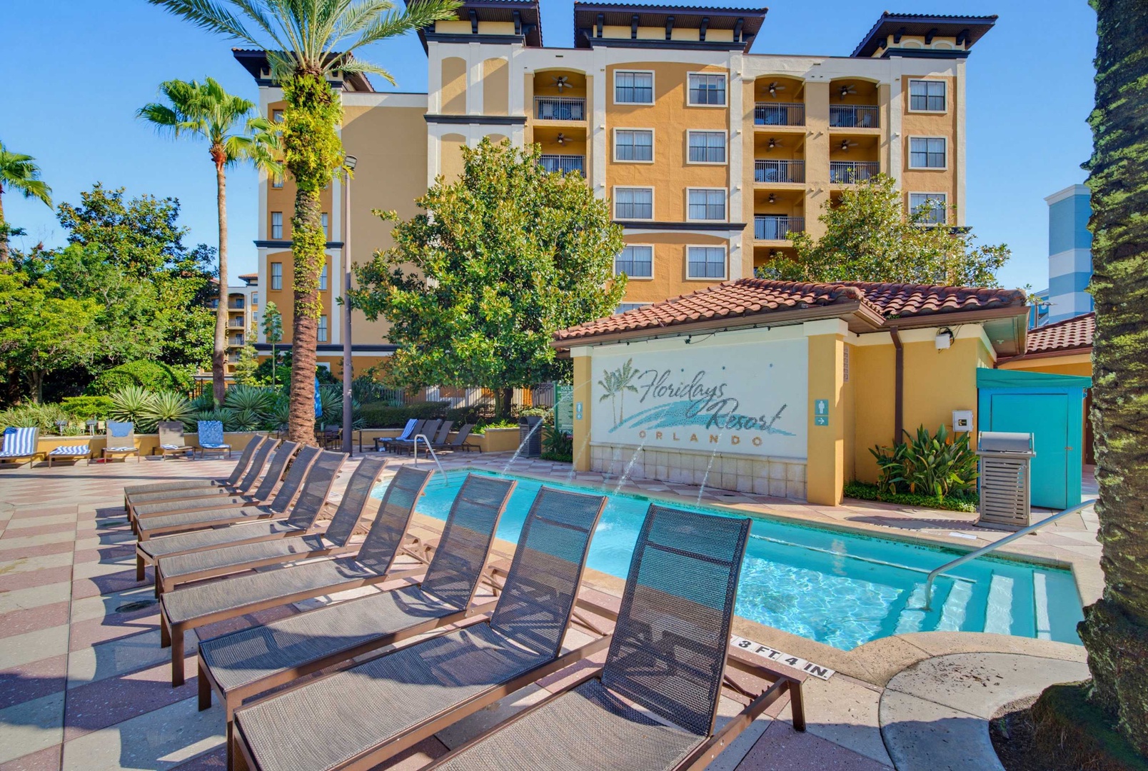 A Mediterranean-style vacation resort with palm trees, loungers by the pool, and balconies overlooking lush tropical gardens under clear blue skies.