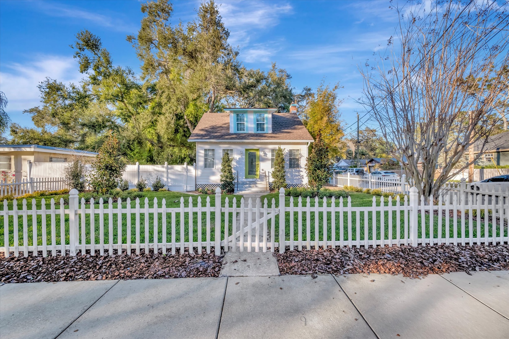 Charming single-story cottage with classic white picket fence and welcoming green door, set in a peaceful neighborhood surrounded by mature trees and manicured landscaping.