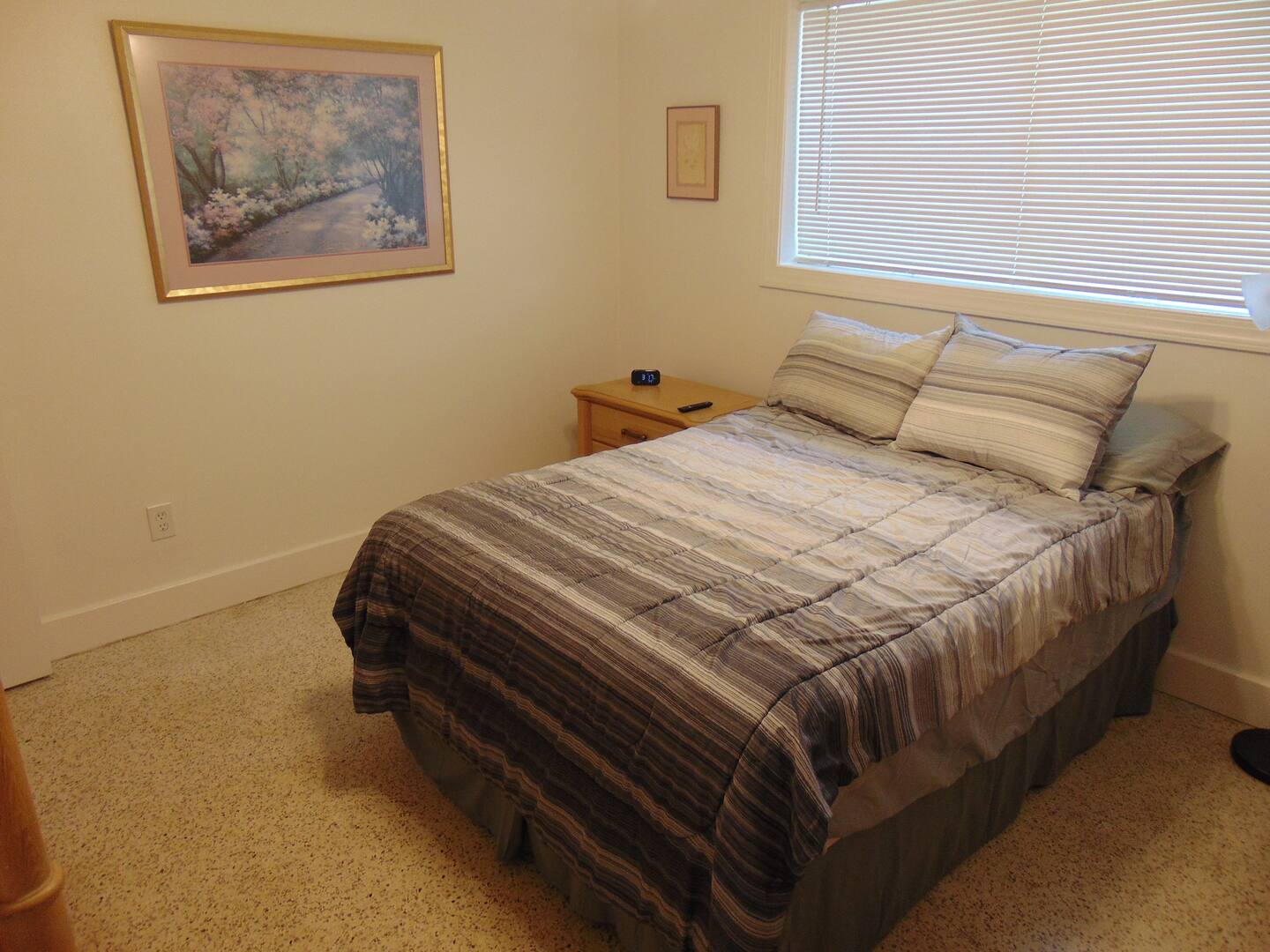 Step into your peaceful bedroom retreat featuring a cozy queen bed dressed in soft striped linens. Natural light filters through the window blinds, creating a warm and restful atmosphere for your stay.