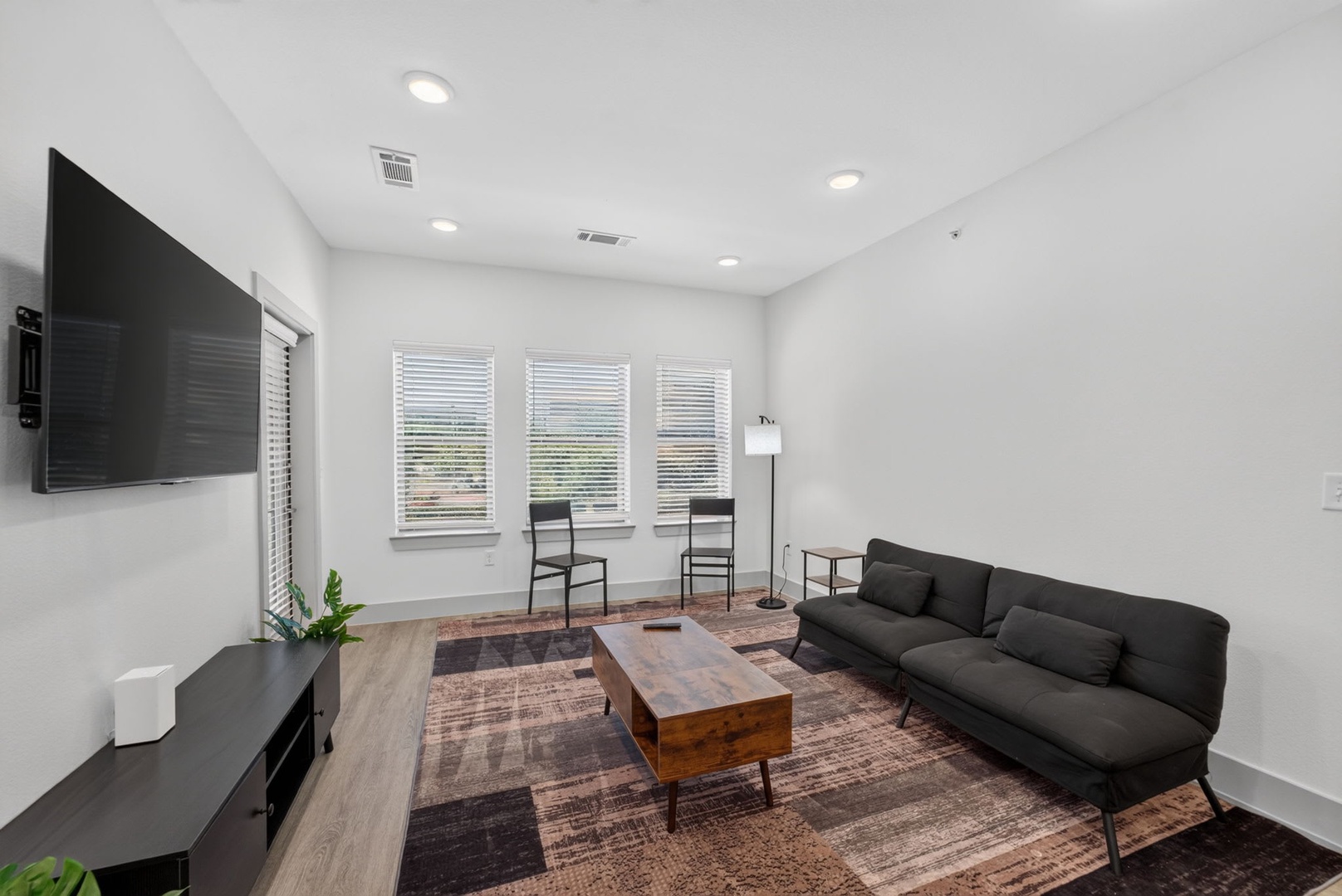 Unwind in your light-filled living room where sleek modern design meets comfort. Natural light streams through three windows, complementing the minimalist furnishings and warm wood accents that create your perfect retreat.