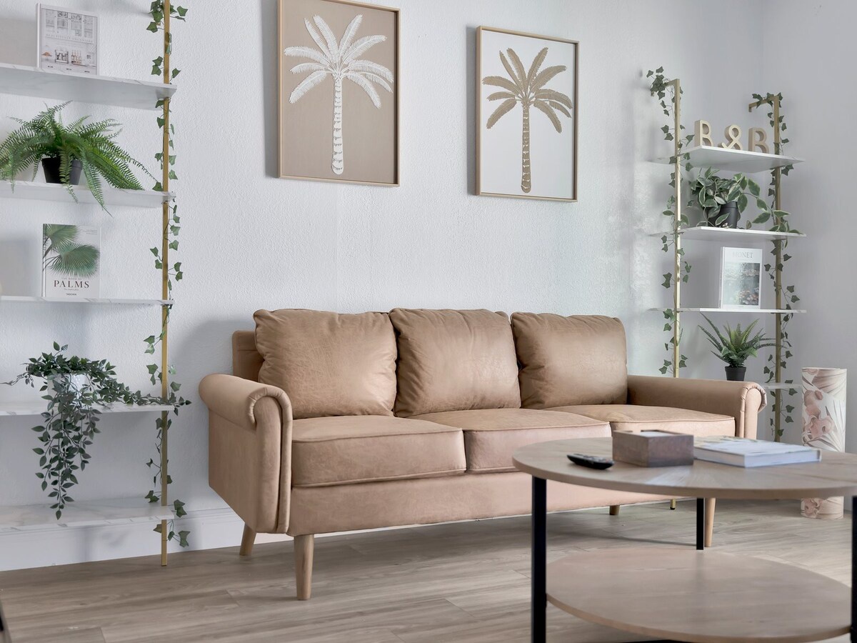 Sink into this plush tan sofa in your airy living space, surrounded by lush greenery and coastal-inspired touches that invite you to unwind.