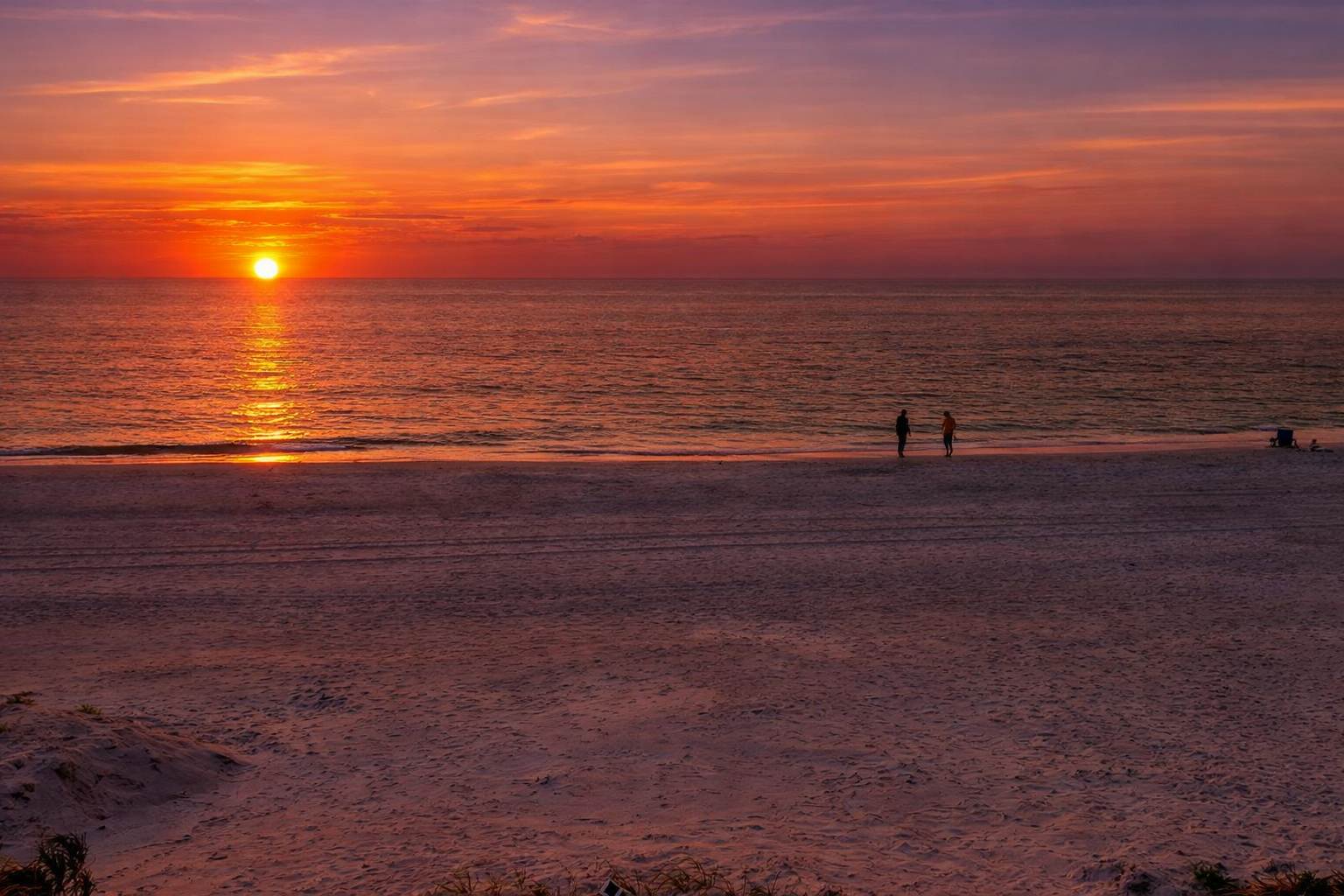 A stunning sunset paints the beach in golden and coral hues, with two silhouettes creating a serene moment at the water's edge—part of the beautiful coastal setting surrounding the property.