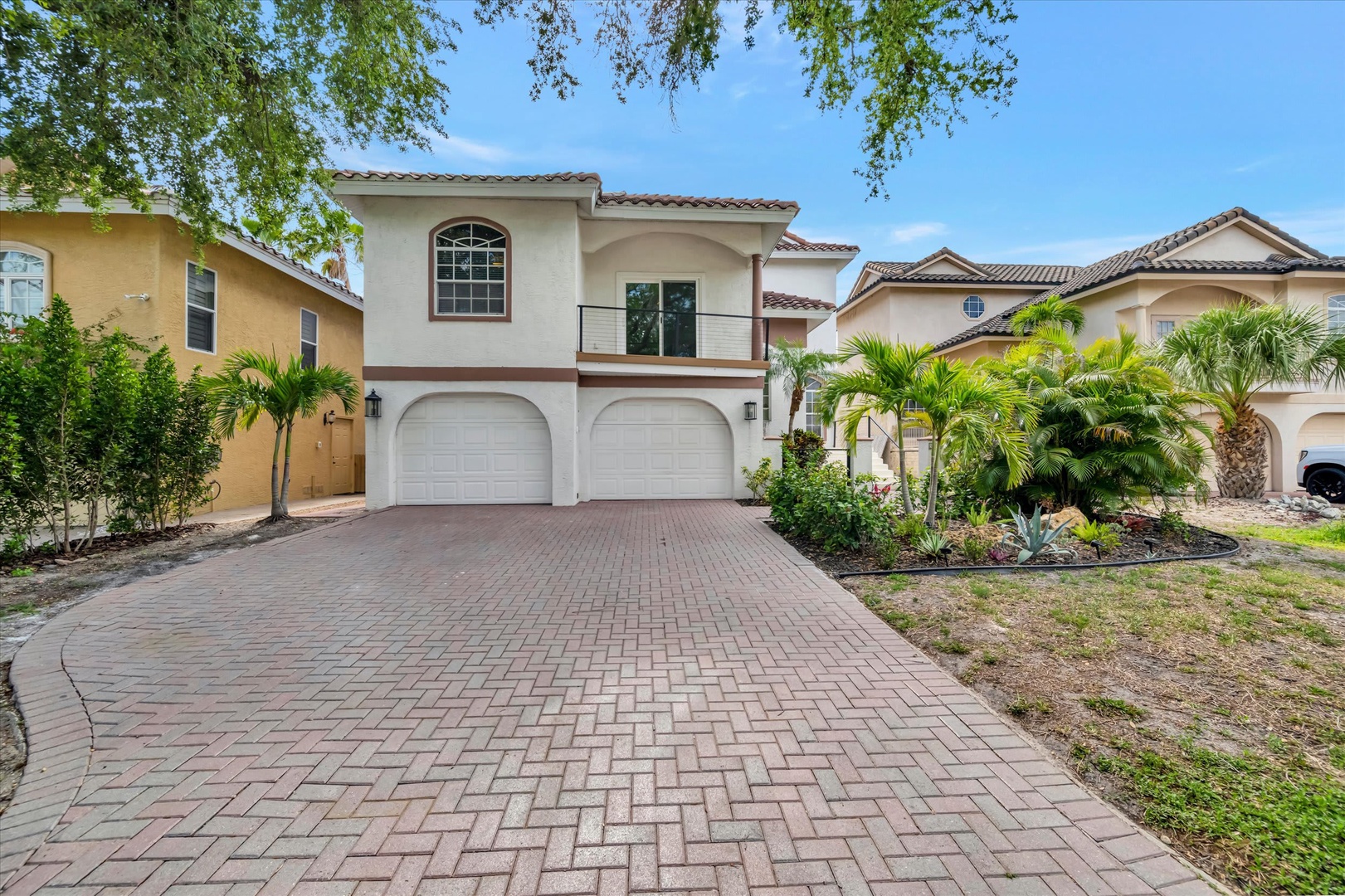 This Mediterranean-style home sits in a quiet residential neighborhood, framed by lush tropical palms and mature landscaping. A brick-paver driveway leads to the two-car garage beneath the main living space.