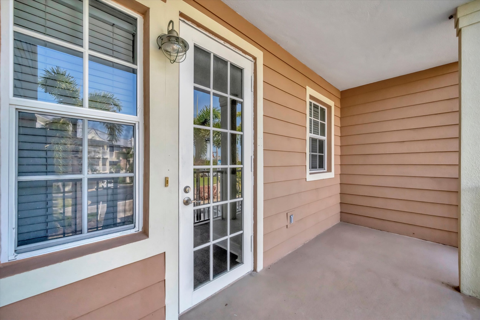 This is the entrance to the property, featuring warm peach-toned siding and a covered entryway with a charming nautical-style light fixture. Palm trees reflect in the windows, setting a tropical, relaxed tone for your stay.