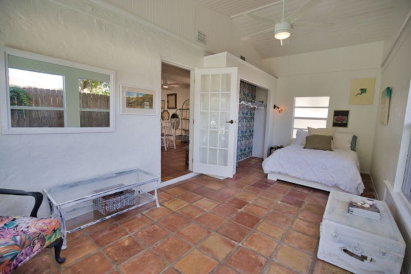 Step into your private retreat featuring charming terracotta floors and natural light through large windows. Your cozy bed awaits, with a glass-paneled door opening to additional living space beyond.
