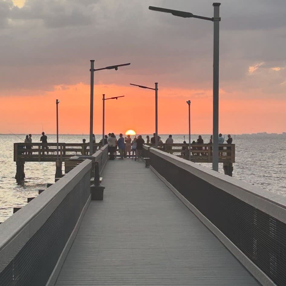 A scenic pier extends into calm waters as visitors gather to watch the sun dip below the horizon, painting the evening sky in warm coral and amber tones.