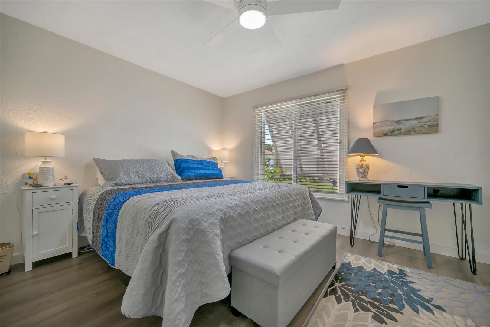 Peaceful bedroom retreat with soft gray tones and ocean blue accents. Natural light streams through plantation shutters, creating a calming coastal vibe. Comfy workspace and plush seating included.