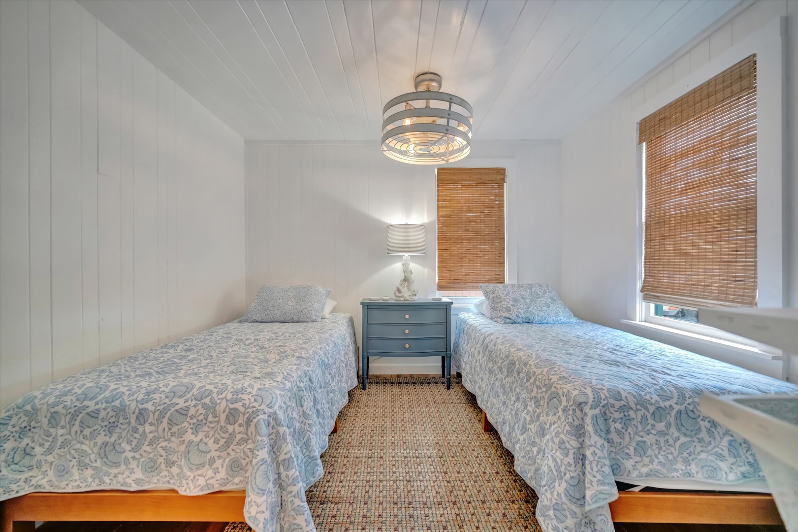 Step into your tranquil coastal retreat with twin beds draped in soft blue linens, a charming nautical-inspired fixture, and natural woven shades filtering gentle sunlight—your peaceful hideaway awaits.