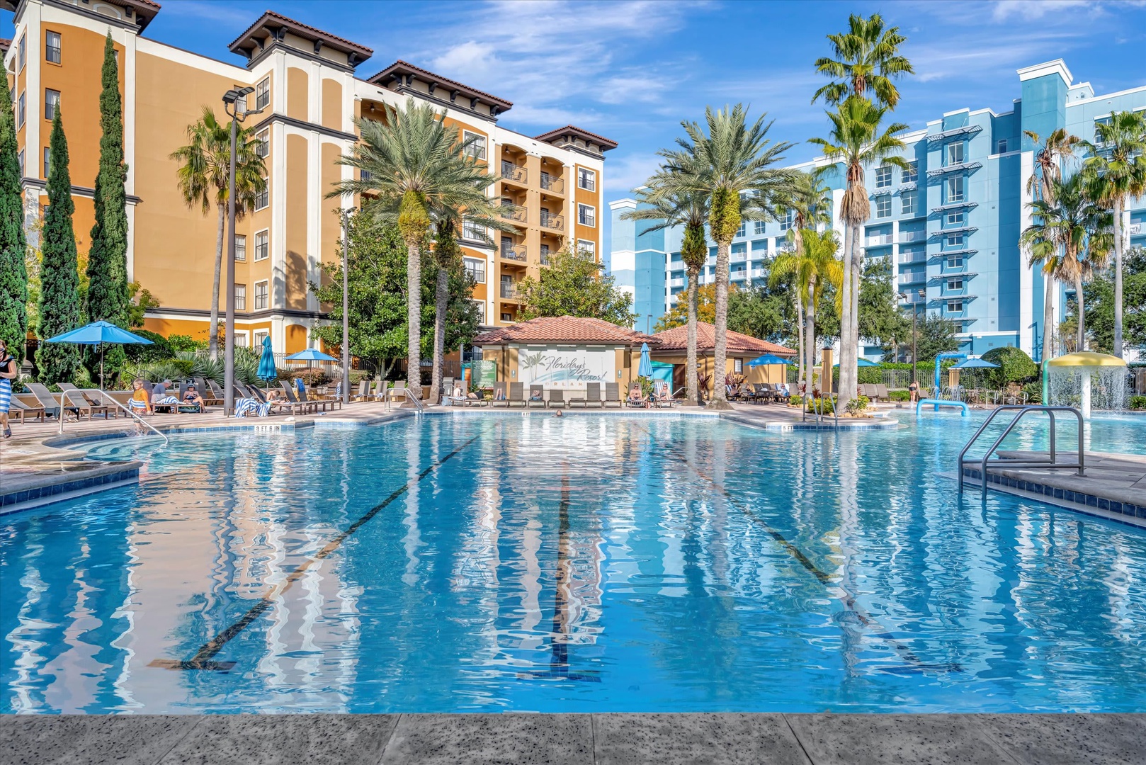 A sparkling outdoor pool surrounded by palm trees and colorful resort buildings under sunny blue skies.