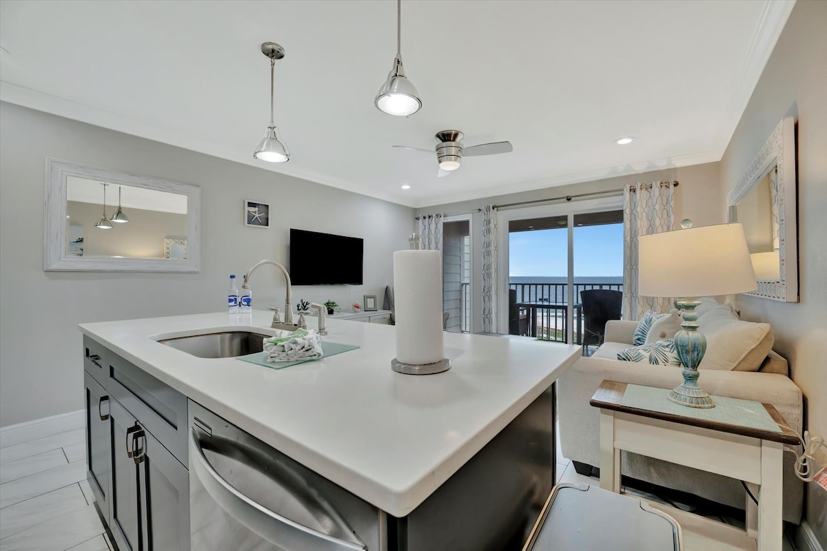 Your inviting open-plan living space features a fully equipped kitchen island with sleek countertops, flowing seamlessly into the living room where coastal breezes drift through glass doors to your private balcony overlooking the ocean.