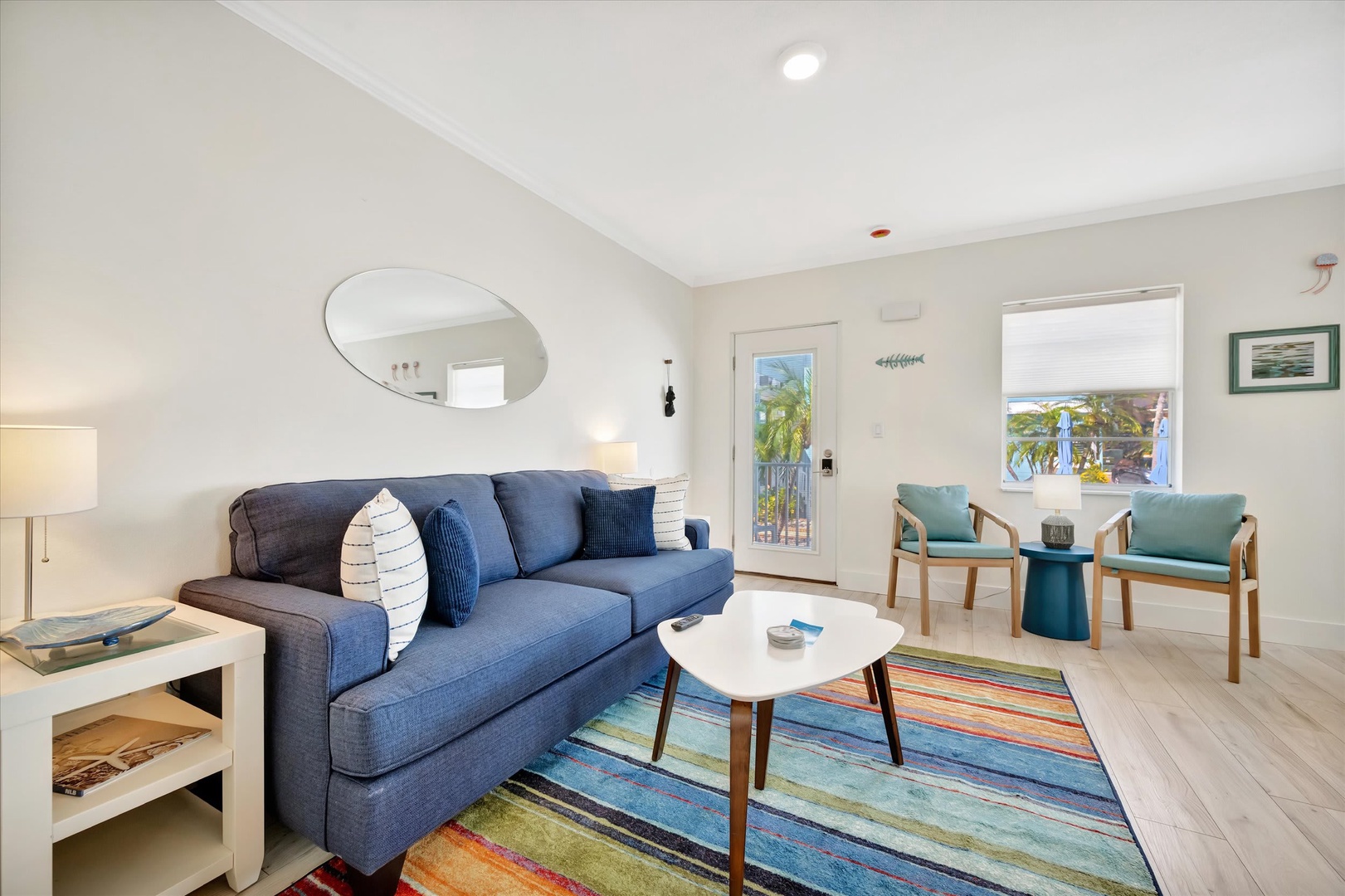 Relax in your bright, coastal-inspired living room with comfortable seating and colorful accents. Through the windows, catch glimpses of palm trees swaying in the breeze—your peaceful retreat awaits.