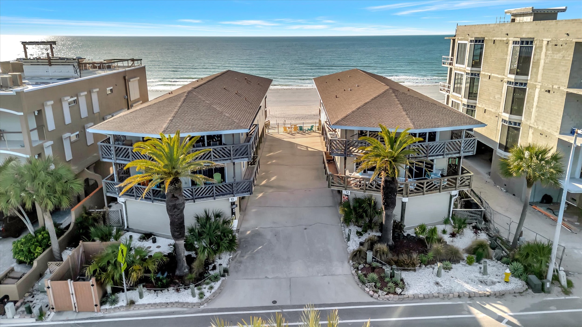 Beachfront property with direct ocean access, surrounded by tropical landscaping and neighboring coastal buildings. The entrance features twin structures with covered balconies and a private walkway leading to the sandy shore.