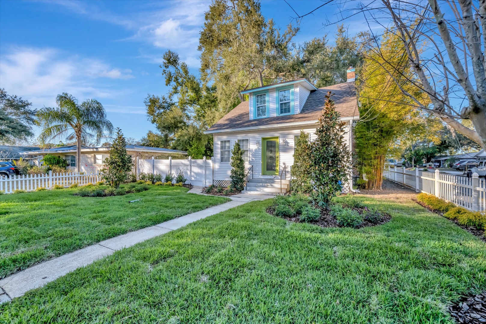 A charming Cape Cod-style cottage nestled in a peaceful neighborhood, featuring a landscaped front lawn, white picket fence, and mature trees providing natural shade throughout the property.