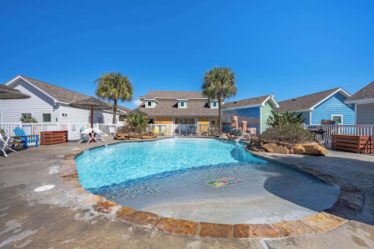 Resort-style pool surrounded by colorful cottages and swaying palms, featuring a zero-entry area perfect for wading and lounging under bright coastal skies.