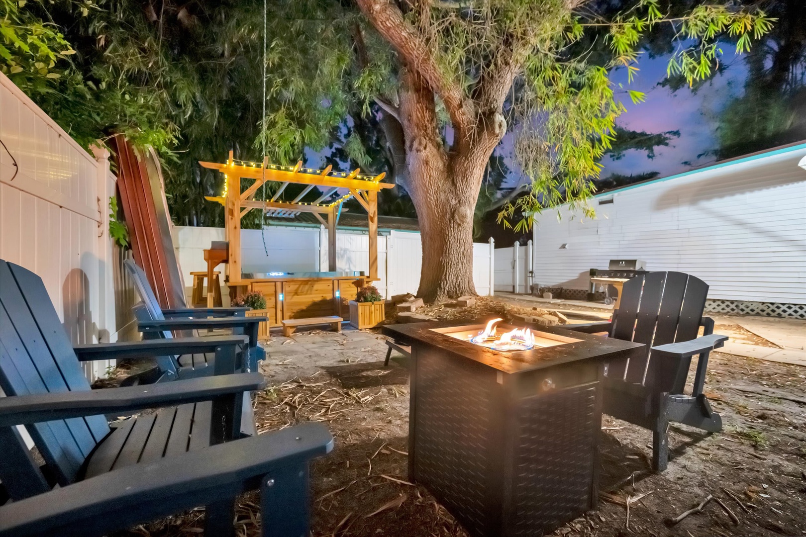 Your private backyard retreat invites you to gather around a glowing fire pit beneath the mature tree, with cozy seating, a charming pergola, and a grill ready for evenings under the stars.